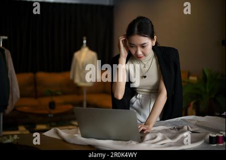 Fokussierte asiatische Modedesignerin, die ihr neues Muster auf ihrem Laptop überprüft, während sie in ihrem Workshop-Studio arbeitet. Stockfoto