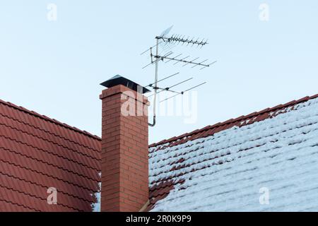 TV-Antenne auf rotem Dach. Fernsehantenne auf dem Dach mit blauem Himmel. Ziegelkamin und schneebedecktes Dach des Hauses und Mediensender, Radio, Radio Stockfoto