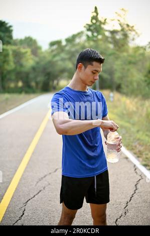 Porträt eines gut aussehenden Asiaten in Sportbekleidung mit einer Flasche Wasserstand auf der Straße in einem Naturpark. Stockfoto