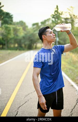 Porträt eines durstigen und erschöpften Asiaten in Sportbekleidung, der sich nach dem Laufen in einem Park Wasser aus einer Flasche ins Gesicht gießt und sich mit Splas erfrischt Stockfoto