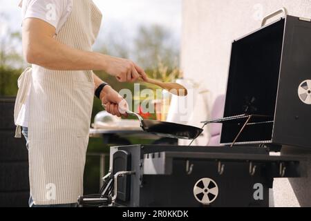 Mittlere Auswahl eines Mannes in der Schürze mit einem Holzlöffel zum Mischen von Gemüse in der Bratpfanne. Bereiten Sie Speisen auf dem Grill zu Stockfoto