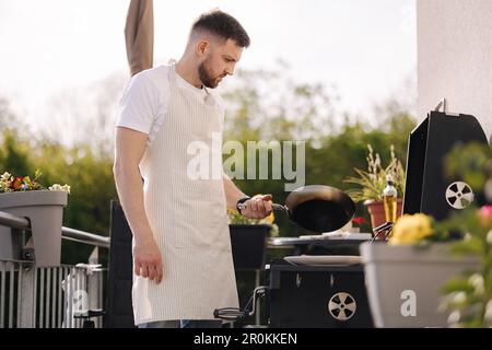 Ein hübscher bärtiger Mann in einer Schürze, der Kartoffeln auf dem Grill brät, indem er eine Karbonstahlpfanne verwendet. Stockfoto