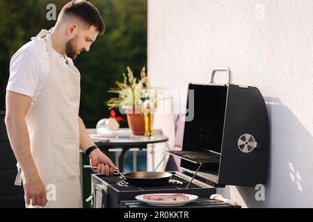 Ein hübscher bärtiger Mann in einer Schürze, der Kartoffeln auf dem Grill brät, indem er eine Karbonstahlpfanne verwendet. Stockfoto