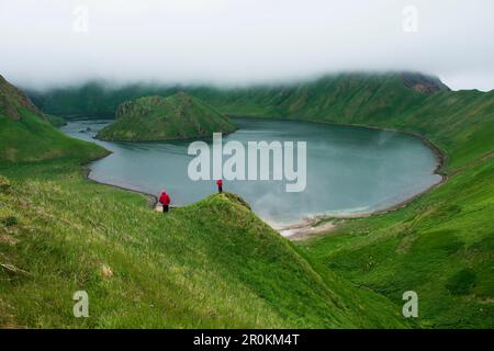 Touristen in roten Parkas stehen auf einer Landzunge vor einer großen wassergefüllten Caldera, Yankicha, Uschischir, Kuril-Inseln, dem Okhotsk-Meer, Russland, Stockfoto