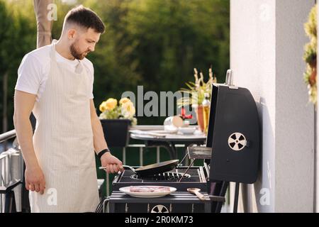 Ein hübscher bärtiger Mann in einer Schürze, der Kartoffeln auf dem Grill brät, indem er eine Karbonstahlpfanne verwendet. Stockfoto