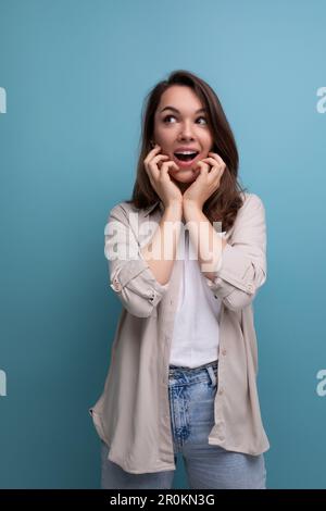 Überraschend emotionale Brünette 30-jährige Frau, die in Hemd und Jeans gekleidet ist, sieht auf blauem Hintergrund weg Stockfoto