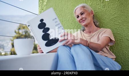 Die einfachen Dinge im Leben. Eine attraktive Seniorin, die eine Tasse Kaffee genießt, während sie draußen auf ihrem Balkon zu Hause ein Buch liest. Stockfoto