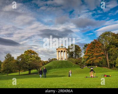 Ein junges Paar, das im Herbst vor den Monopteros im Englischen Garten Yoga auf dem Gras praktiziert, Passanten, die über das Grün, München, spazieren Stockfoto