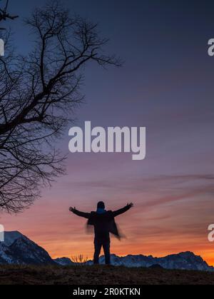 Zur blauen Stunde steht ein Mann auf einem Hügel neben einem Baum mit ausgestreckten Armen und blickt auf das Karwendel- und Wetterstein-Gebirge Esch Stockfoto