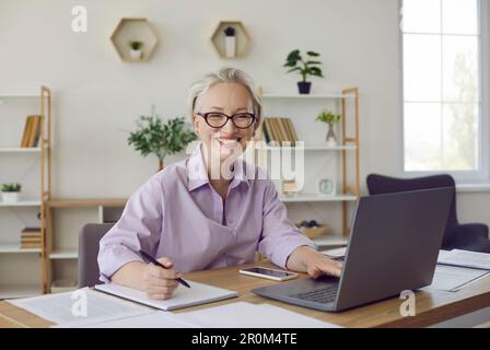 Glückliche Geschäftsfrau, die am Schreibtisch sitzt, an einem Laptop arbeitet und sich Notizen in einem Notizbuch macht Stockfoto