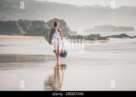 Eine Frau mit einem Surfbrett an der Küste Nordspaniens geht am Strand entlang Stockfoto