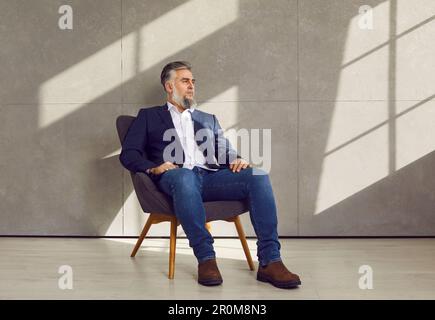 Porträt eines erfolgreichen, selbstbewussten Geschäftsmannes, der an der grauen Bürowand auf einem Stuhl sitzt Stockfoto
