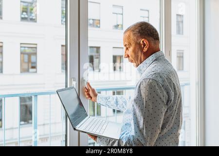 Attraktiver Mann im Hemd steht neben dem Fenster mit Laptop Stockfoto