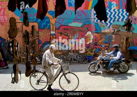 Ein erfahrener Fahrradfahrer trifft auf einem Motorrad vor einer hell bemalten Mauer in der marokkanischen Altstadt Marrakesch auf Touristen Stockfoto