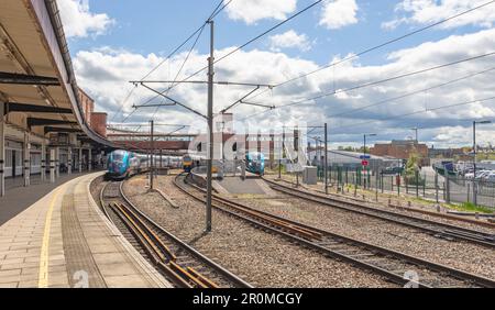 Drei Züge warten am Bahnhof. Eine Fußgängerbrücke verbindet die Plattformen, und ein Himmel mit Wolken befindet sich darüber. Ein Parkplatz befindet sich auf einer Seite. Stockfoto