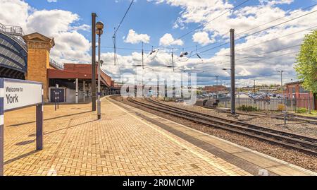 Züge warten am Bahnhof. Eine Fußgängerbrücke verbindet die Plattformen, und ein Himmel mit Wolken befindet sich darüber. Ein Begrüßungsschild ist im Vordergrund und ein Auto Pa Stockfoto