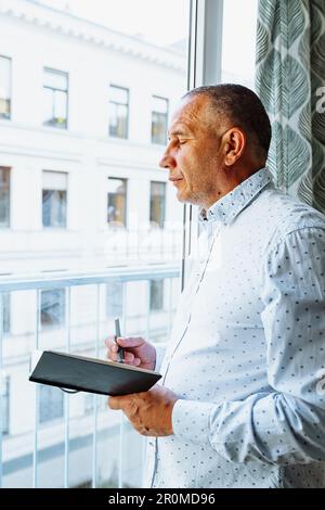 Attraktiver Mann im Hemd steht neben dem Fenster mit Notizblock Stockfoto