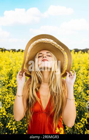 Porträt einer jungen, wunderschönen blonden Frau, die ein rotes Kleid und einen Hut trägt, inmitten eines Feldes blühender gelber Rapsblumen Stockfoto