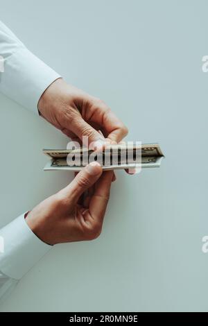 Ein Mann, der ein weißes Langarmhemd trägt und Dollarnoten zählt, sitzt an einem weißen Tisch Stockfoto