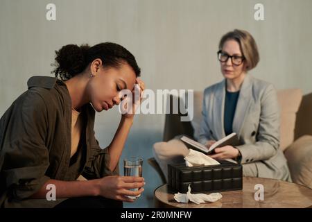 Traurige junge afroamerikanische Patientin einer Beraterin mit einem Glas Wasser in der Hand, die ihr Problem einer Psychotherapeutin in der Praxis beschreibt Stockfoto