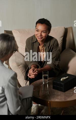 Glückliche junge afroamerikanische Patientin, die in einem bequemen Sessel vor der Psychologin sitzt und sie während der Sitzung ansieht Stockfoto