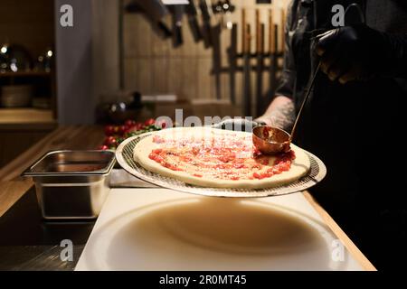 Nahaufnahme eines professionellen Küchenchefs, der die Mischung der Zutaten auf der Oberfläche des Fladenbrots auf einem Metalltablett verteilt, bevor er es zum Backen in den Ofen stellt Stockfoto