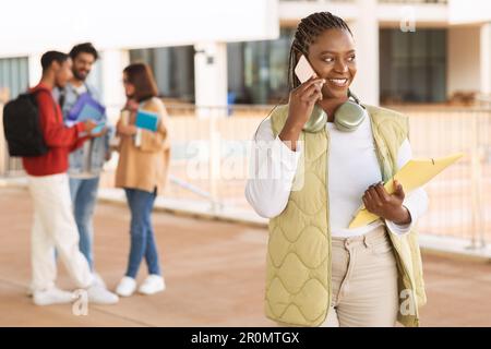 Positive schwarze Studentin, die gelegentlich telefoniert, bereit für Unterricht und Test an der Universität Stockfoto