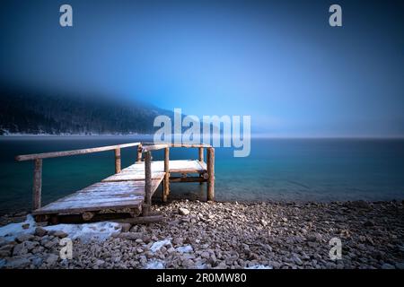 Lichtstrahl auf schneebedeckten Holzsteg am Walchensee im Nebel im Winter, lange Exposition, Alpenausläufer, Bayern Deutschland Stockfoto