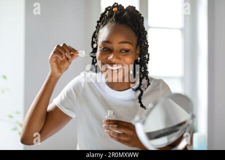 Tägliche Hautentgiftungsroutine. Wunderschöne schwarze Dame, die zu Hause vor dem Spiegel sitzt und ein feuchtigkeitsspendendes Serum aufträgt Stockfoto