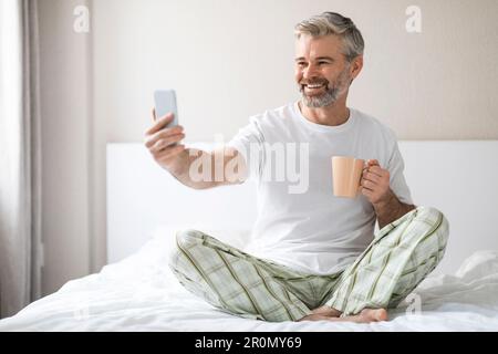 Glücklicher Mann, der im Bett telefoniert und Kaffee trinkt Stockfoto