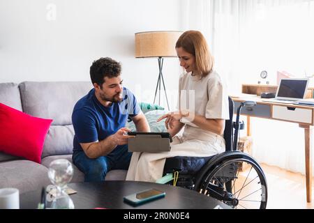 Bärtiger Mann sitzt auf dem Sofa mit Frau im Rollstuhl sitzt auf Rollstuhl in lässiger Kleidung und diskutiert und sieht sich Videos auf einem Tablet an Stockfoto