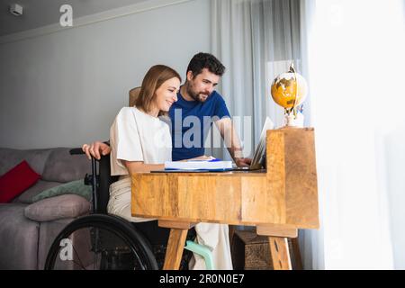 Liebenswerter, liebevoller Ehemann, der seiner Frau im Rollstuhl hilft, mit einem Büroprojekt zu Hause, das mit dem Laptop funktioniert Stockfoto