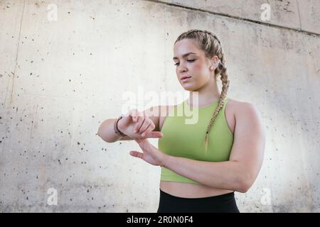Beruhigen Sie junge Sportlerinnen in Sportbekleidung, die neben Betonwänden stehen und nach unten schauen, während sie beim Fitnesstraining eine moderne Smartwatch verwenden Stockfoto