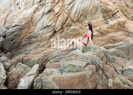 Von unten sehen Sie die fröhliche junge Touristin in der Bikinizone und Sonnenbrille, die glücklich lächelt, während sie sich während des Sommerabenteuers auf einem rauen felsigen Hügel ausruhen Stockfoto