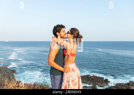 Seitenblick auf ein fröhliches junges Paar im Sommer, stehen an der felsigen Küste und umarmen, während sie lächeln und den Urlaub zusammen gegen das winkende Meer genießen Stockfoto