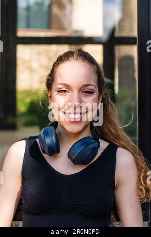 Fröhliche junge Frau mit schnurlosem Headset und Aktivkleidung, die fröhlich lächelt und vor verschwommenem Hintergrund in die Kamera schaut Stockfoto