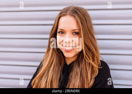 Porträt einer positiven jungen Frau mit langen dunklen Haaren, die lächelt und in die Kamera schaut, während sie gegen die weiße Wand steht Stockfoto