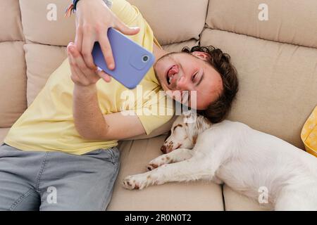 Von oben: Lächelnder junger Transgender-Mann in gelbem Hemd, der auf dem Sofa sitzt und Selbstporträt mit weißem schlafenden Hund macht, während er Zeit verbringt Stockfoto
