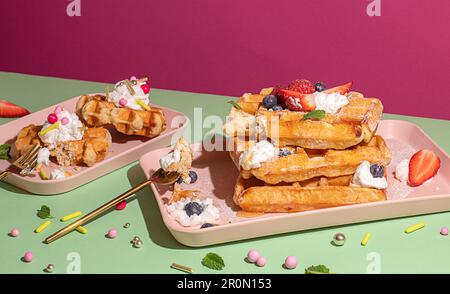 Von oben schmackhafte süße Waffeln garniert mit Beerenobstsauce und Sahne serviert auf einem rosa Tablett auf einem farbenfrohen Tischhintergrund in einer hellen Küche Stockfoto