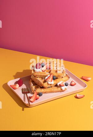 Von oben schmackhafte süße Waffeln garniert mit Beerenobstsauce und Sahne serviert auf einem rosa Tablett auf einem farbenfrohen Tischhintergrund in einer hellen Küche Stockfoto