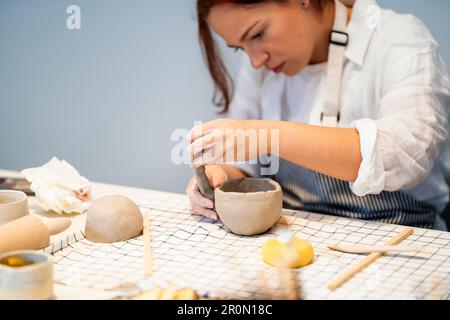 Fokussierte junge Kunsthandwerkerin in weißem Hemd und Schürze, die eine Tonschale bildet und mit verschiedenen Werkzeugen handgefertigte Keramik herstellt Stockfoto