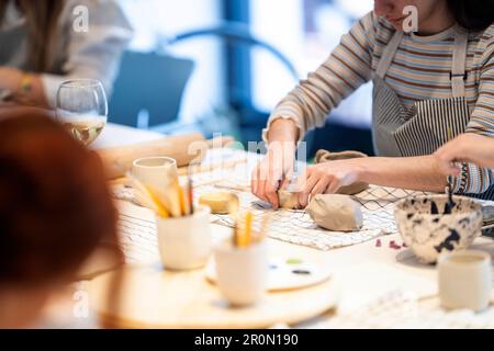 Unbekannte Künstlerinnen in Schürzen, die am Tisch sitzen und handgemachte Tonbecher herstellen, während sie sich während des Unterrichts in einer modernen Werkstatt versammeln Stockfoto