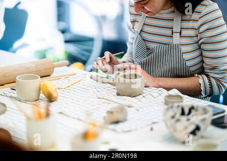 Pflanzkonzentriertes junges Weibchen in der Schürze, das Keramikbecher herstellt, während es mit einem professionellen scharfen Werkzeug in der Werkstatt auf Lehm verziert Stockfoto