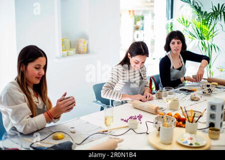 Gruppe positiver junger Künstlerinnen in Schürzen, die sich am Tisch mit verschiedenen Gerichten und Getränken versammeln, während sie Töpferunterricht zusammen in moderner technik machen Stockfoto