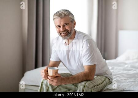 Glücklicher Mann mittleren Alters, der auf dem Bett sitzt und Kaffee trinkt Stockfoto