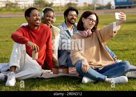 Glückliche internationale Millennials Studenten lernen, trinken Kaffee, entspannen, auf Gras sitzen, Selfie machen Stockfoto
