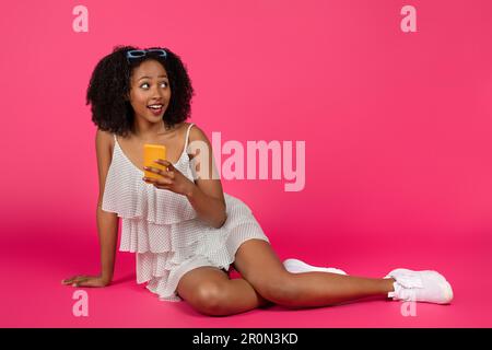 Lächelnde junge afroamerikanische, lockige Frau im Kleid, die sich mit dem Smartphone unterhält, auf dem Boden sitzt Stockfoto