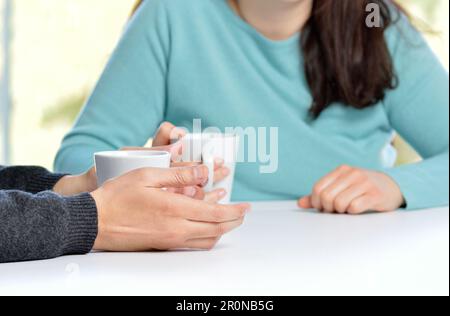 Profil mit Hintergrundbeleuchtung von Händen zweier Freunde oder Paare, die sich unterhalten und Kaffeetassen halten, die zu Hause auf einem Tisch sitzen und ein Fenster im Hintergrund haben Stockfoto