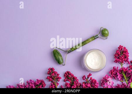 Cremefarbenes Glas, Walze mit Jadegesicht und lila Blumen auf lila Hintergrund. Naturkosmetik, Hautpflegekonzept. Draufsicht, flach liegend, Kopierbereich. Stockfoto