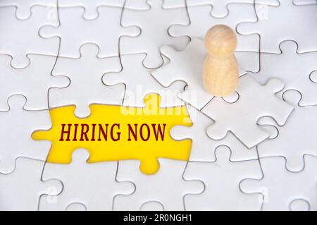 „Hiring NOW“-Text zu fehlendem Puzzle mit Holzfigur-Hintergrund. Beschäftigungskonzept. Stockfoto